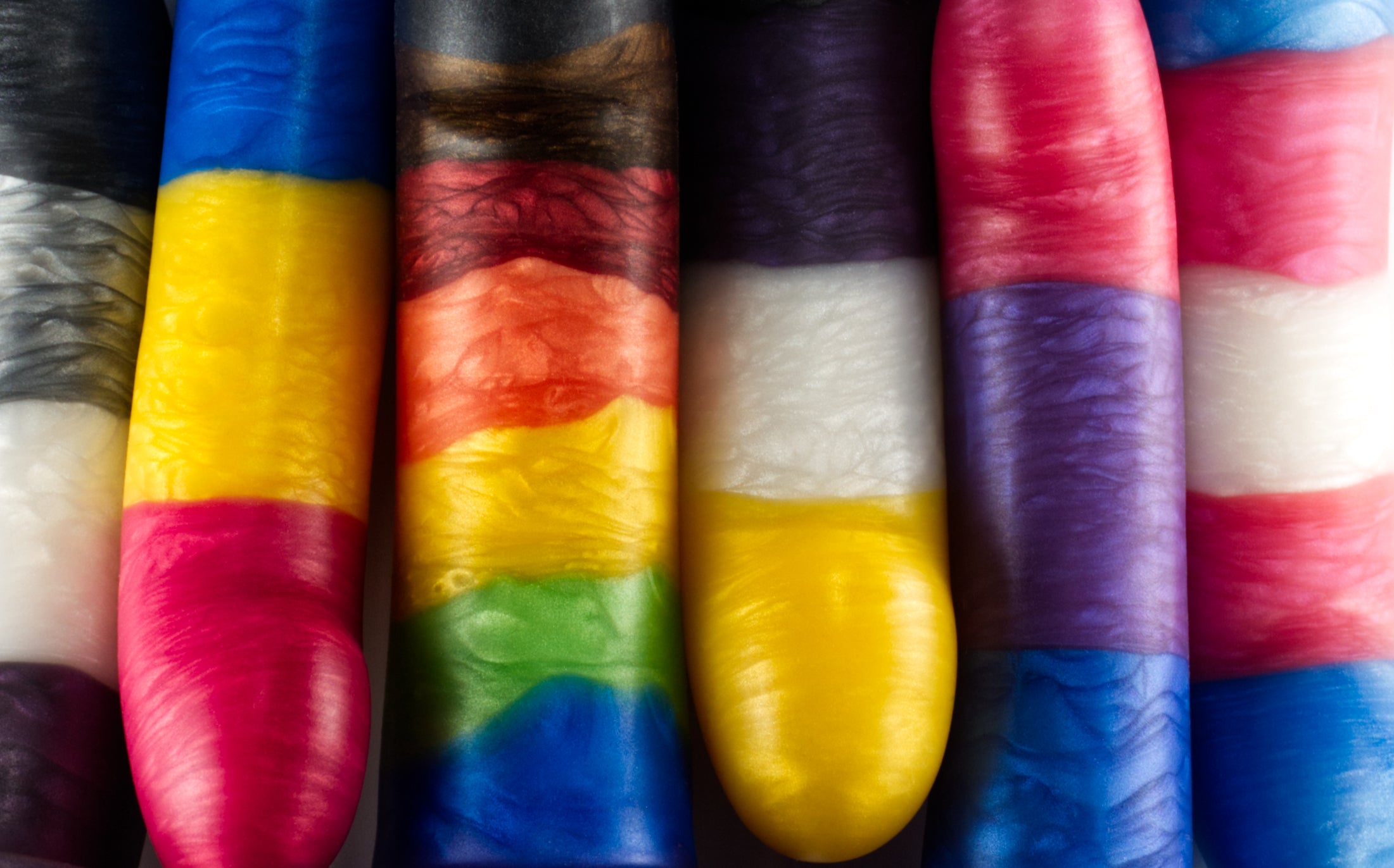 Top view of a row of Sidekick dildos, each poured in a different Pride flag striped color.