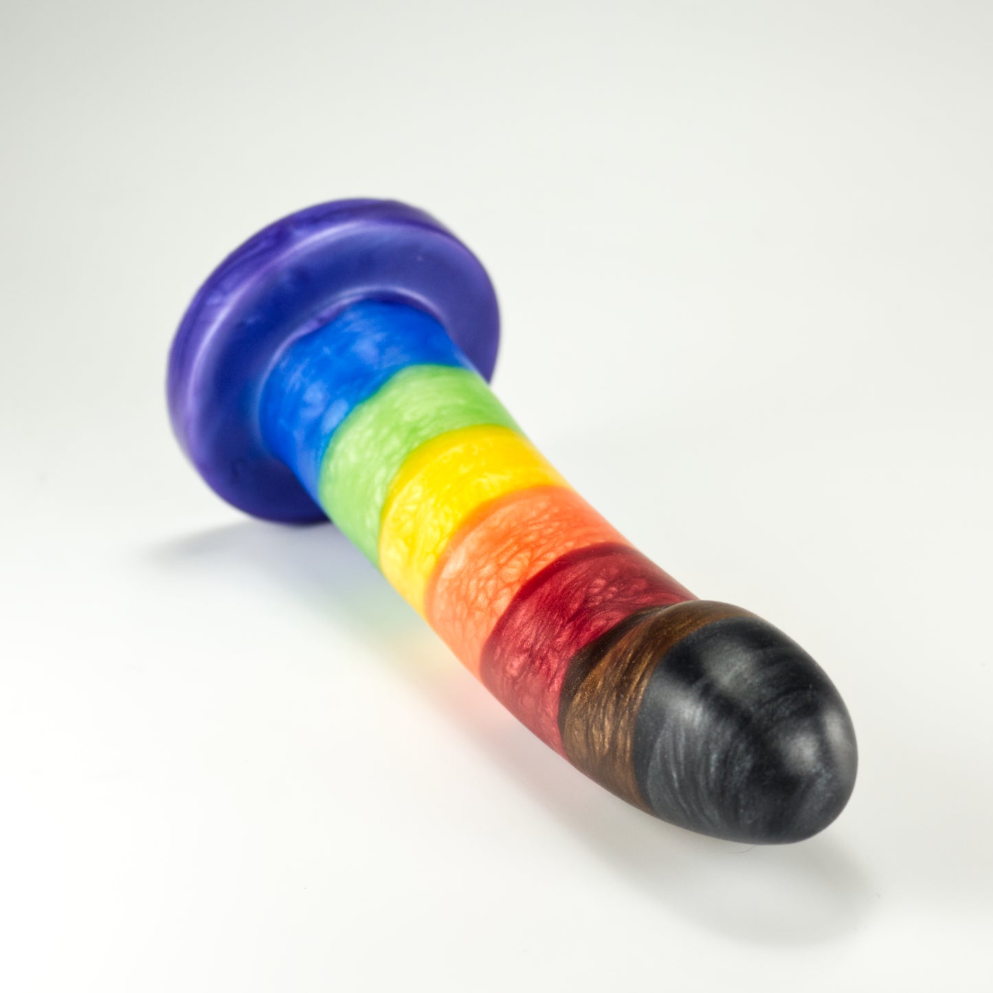 Top view of the Sidekick Small, a dildo with a smooth cylinder shaped body, and a fingertip-shaped tip, and a pronounced cliff shape where they meet. Color is rainbow striped with additional black and brown stripes at tip.
