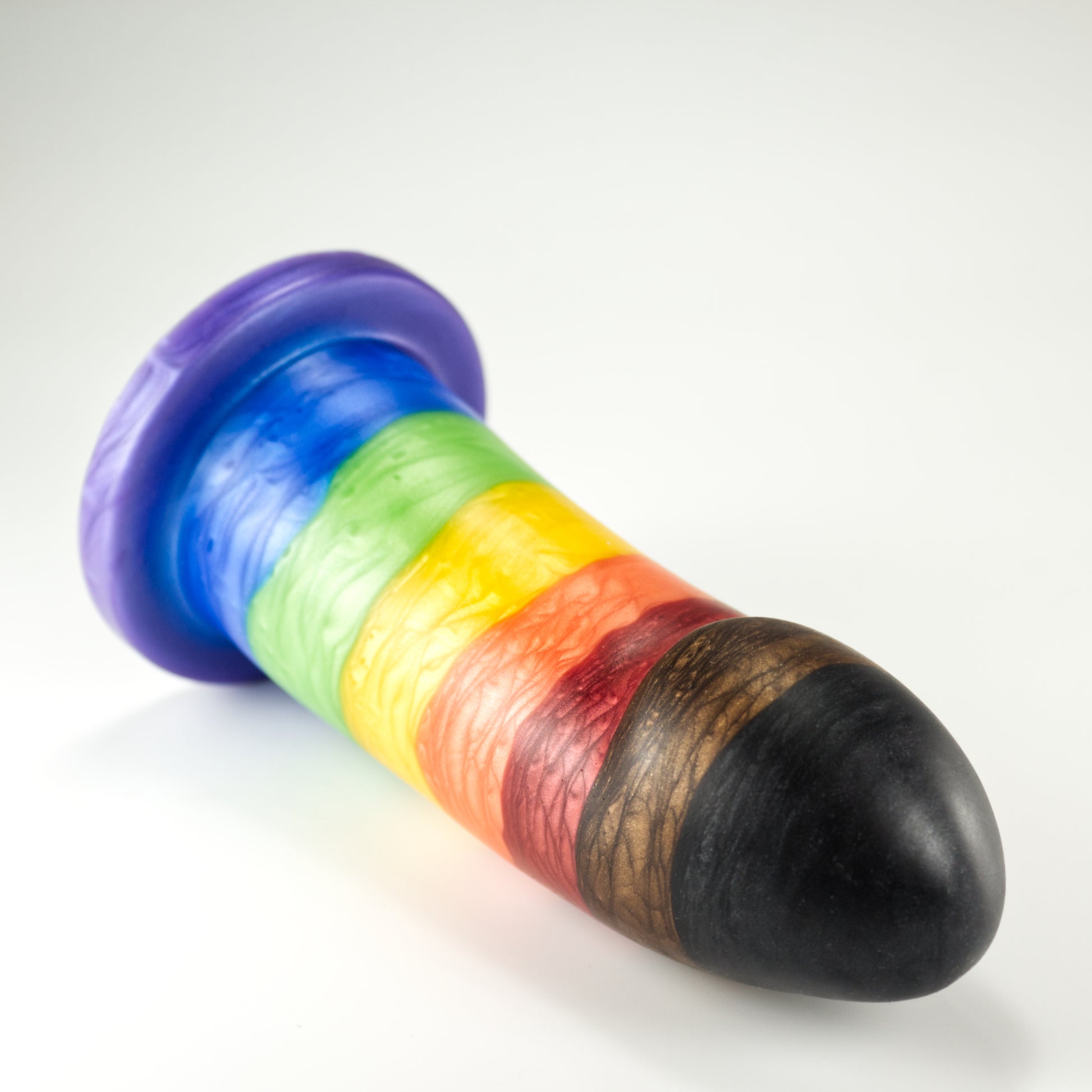 Top view of the Sidekick Extra Large, a dildo with a smooth cylinder shaped body, and a fingertip-shaped tip, and a pronounced cliff shape where they meet. The color is rainbow stripe with additional black and brown stripes at tip.